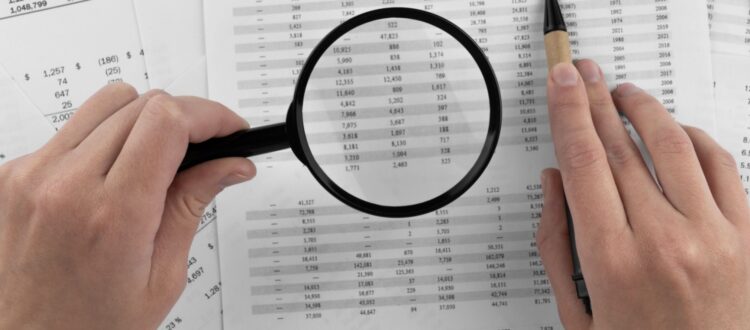 Somebody holding a magnifying glass to paperwork covered in numbers.