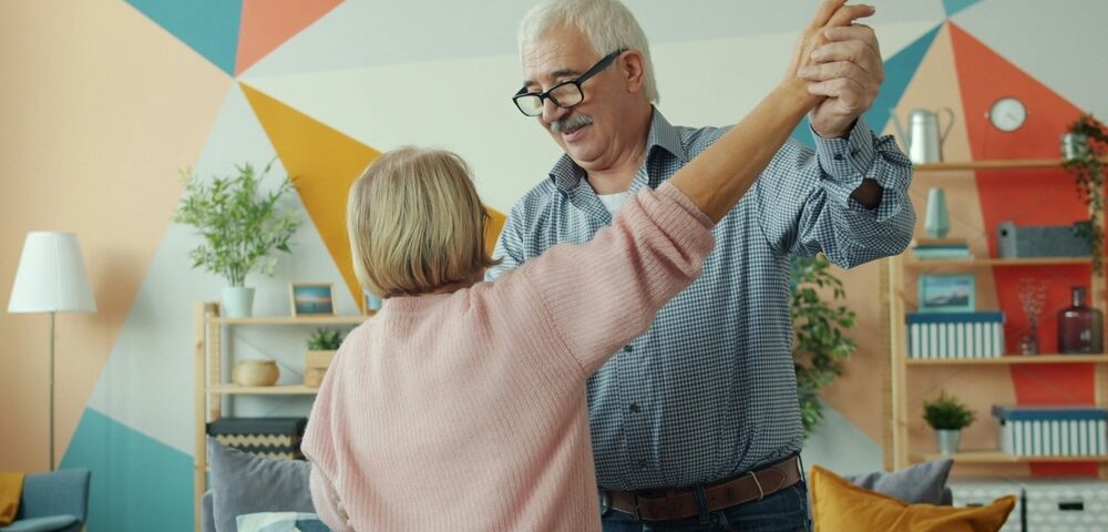 Two happy pensioners dancing at home, holding hands and having fun